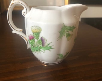 May include: A white porcelain creamer with a gold trim and a floral design featuring green leaves and purple and yellow thistles.