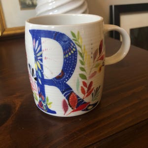 K&ouml;nnte beinhalten: Eine wei&szlig;e Keramiktasse mit einem blauen Buchstaben "R", umgeben von bunten Blumenmustern. Die Tasse hat einen Henkel und ist perfekt f&uuml;r ein hei&szlig;es Getr&auml;nk.