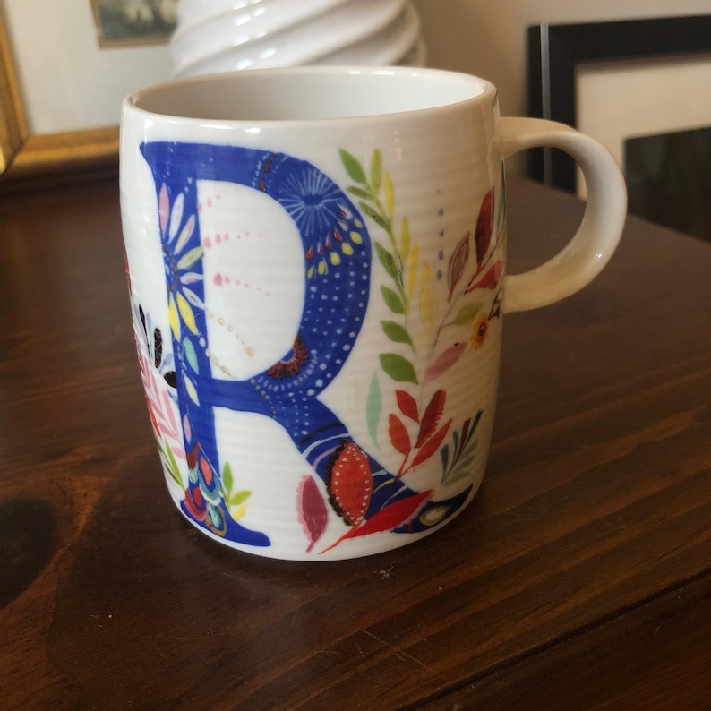 K&ouml;nnte beinhalten: Eine wei&szlig;e Keramiktasse mit einem blauen Buchstaben "R", umgeben von bunten Blumenmustern. Die Tasse hat einen Henkel und ist perfekt f&uuml;r ein hei&szlig;es Getr&auml;nk.