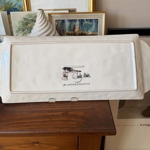 May include: A rectangular, off-white serving platter with raised edges and handles. The bottom of the platter features a small illustration and the text "Sally Muir for Anthropologie."