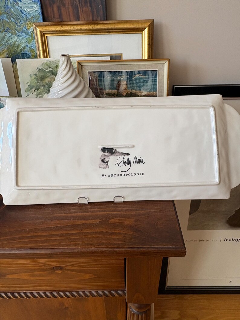 May include: A rectangular, off-white serving platter with raised edges and handles. The bottom of the platter features a small illustration and the text "Sally Muir for Anthropologie."