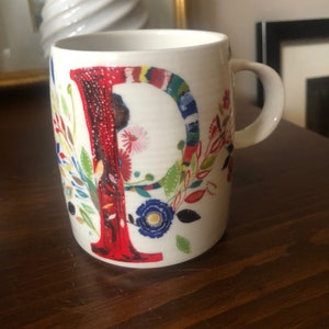 K&ouml;nnte beinhalten: Wei&szlig;e Keramiktasse mit einem roten Buchstaben "P", umgeben von bunten Blumenmustern. Die Tasse hat einen Henkel.