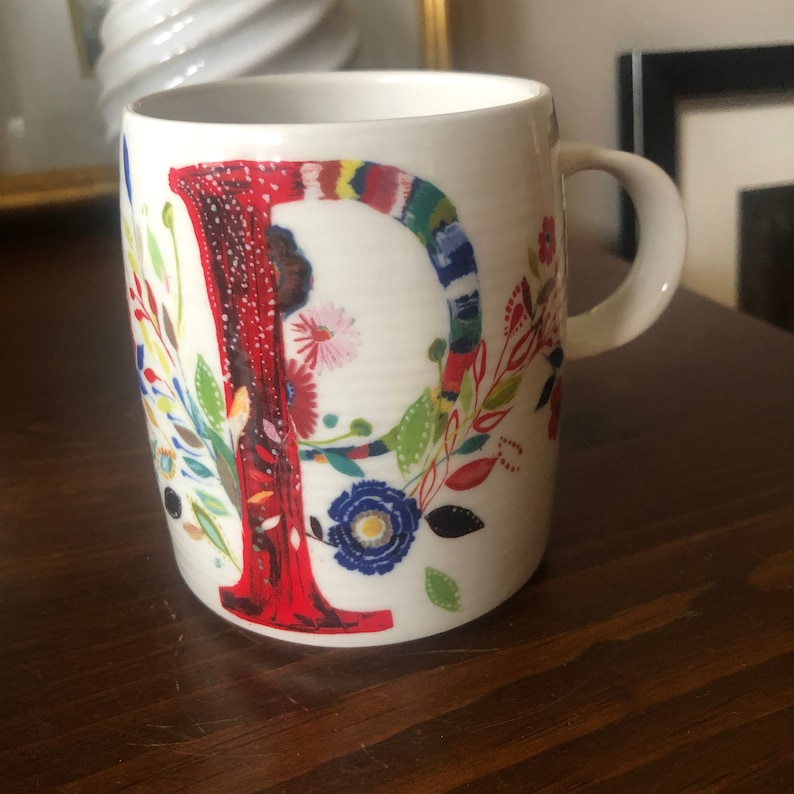K&ouml;nnte beinhalten: Wei&szlig;e Keramiktasse mit einem roten Buchstaben "P", umgeben von bunten Blumenmustern. Die Tasse hat einen Henkel.