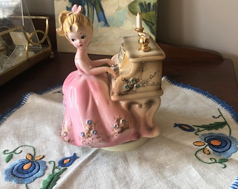 Może przedstawiać: Antyczna figurka porcelanowa przedstawiająca dziewczynkę grającą na pianinie. Dziewczynka ma na sobie różową sukienkę z złotymi akcentami i siedzi przy białym pianinie ze złotymi akcentami. Pianino ma świeczkę na górze.