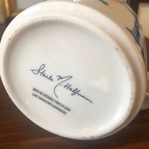 K&ouml;nnte beinhalten: Wei&szlig;e Keramiktasse mit blau-wei&szlig;em Blumenmuster. Am Boden der Tasse befindet sich die Signatur "Carla M. Hallman" und der Text "Dishwasher & Microwave Safe Made in China / Fabrique en Chine".