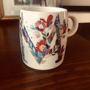 K&ouml;nnte beinhalten: Wei&szlig;e Keramiktasse mit einem Blumendesign, das einen gro&szlig;en Buchstaben "M" in der Mitte zeigt. Die Blumen sind in Rot-, Blau-, Gr&uuml;n- und Gelbt&ouml;nen bemalt.