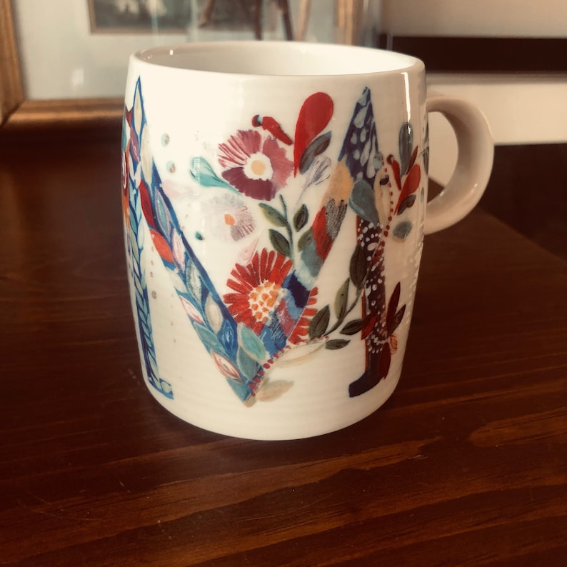 K&ouml;nnte beinhalten: Wei&szlig;e Keramiktasse mit einem Blumendesign, das einen gro&szlig;en Buchstaben "M" in der Mitte zeigt. Die Blumen sind in Rot-, Blau-, Gr&uuml;n- und Gelbt&ouml;nen bemalt.