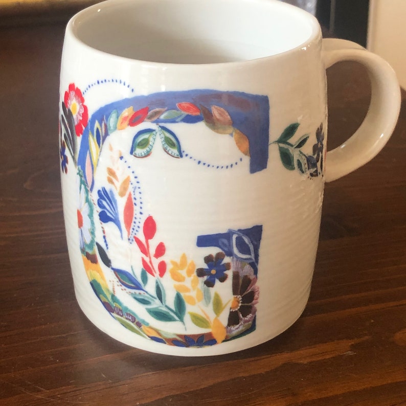 K&ouml;nnte beinhalten: Wei&szlig;e Keramiktasse mit einem blauen und floralen Design mit dem Buchstaben "G". Das Blumenmuster umfasst die Farben Rot, Gelb, Blau, Gr&uuml;n und Braun.