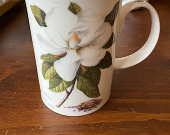 May include: White ceramic mug featuring a detailed illustration of a magnolia flower with green leaves and a brown stem. The mug has a curved handle and the botanical name, Magnolia, is printed on the side. The mug is set on a wooden surface.