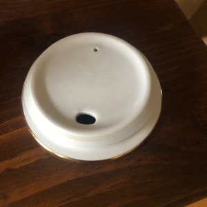 Op de afbeelding: Een wit keramisch deksel met een gouden rand en een klein gaatje in het midden. Het deksel is ontworpen voor een koffiemok of theekopje.