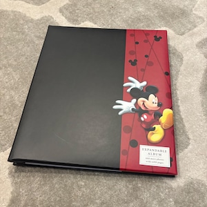 May include: Black expandable photo album with a red spine featuring Mickey Mouse. The album has a black cover and a red spine with Mickey Mouse graphics and the text "EXPANDABLE ALBUM".