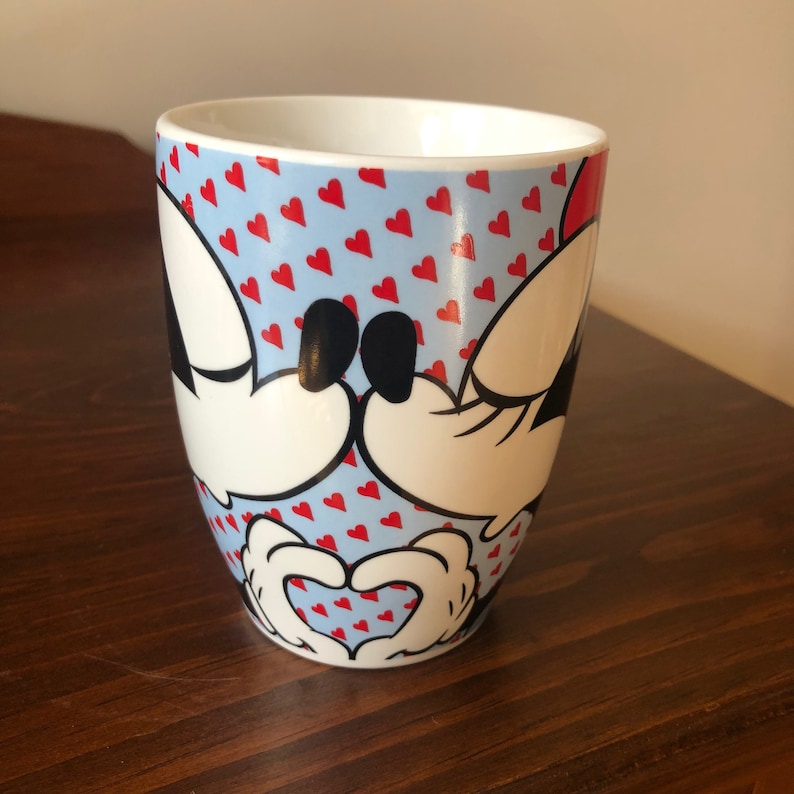 Pu&ograve; includere: Una tazza in ceramica bianca con sfondo blu e cuori rossi. Topolino e Minnie Mouse si tengono per mano a forma di cuore.
