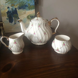 May include: A white teapot, milk jug and sugar bowl with a gold trim and pink rose pattern. The teapot has a lid and a handle. The milk jug and sugar bowl have handles. The set is on a wooden surface.