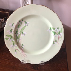 May include: A light green ceramic dinner plate with a gold rim and a white floral design of snowdrops.