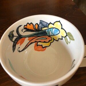 Pu&ograve; includere: Tazza da t&egrave; in ceramica bianca con un design floreale colorato con accenti arancioni, gialli, blu, verdi e neri.