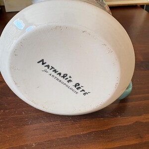 May include: A white ceramic vase with a floral design and a turquoise handle. The bottom of the vase has the text "Nathalie Lete for Anthropologie" in black script.