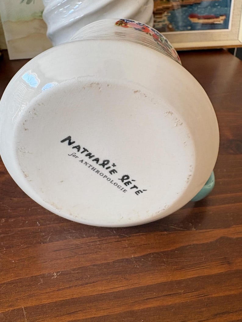 May include: A white ceramic vase with a floral design and a turquoise handle. The bottom of the vase has the text "Nathalie Lete for Anthropologie" in black script.