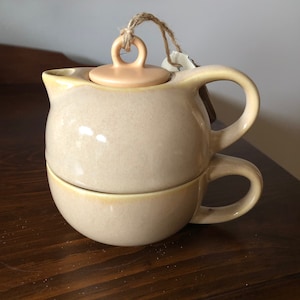 May include: A beige ceramic teapot and cup set with a light brown lid. The teapot has a round body and a handle on the side. The cup has a handle on the side. The set is on a wooden surface.