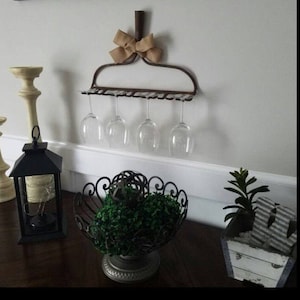 May include: A rustic metal wine glass rack shaped like a rake with a burlap bow. The rack is mounted on a white wall and holds five wine glasses.