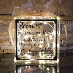 May include: A glass block with a black frame and a white ribbon on top. The block contains a string of white lights and a black vinyl decal with the text "Because someone we love is in Heaven... there's a little bit of HEAVEN in our home."