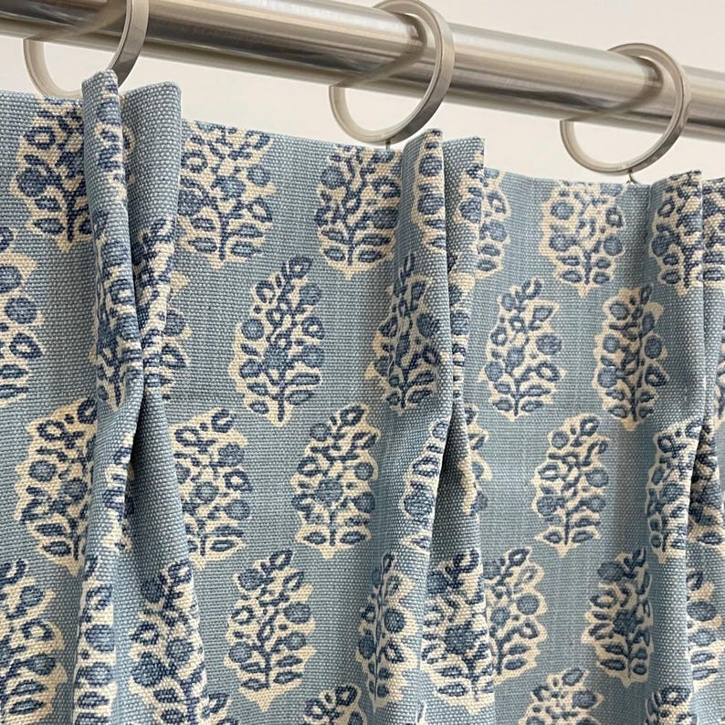 May include: Close-up of blue curtains with a repeating floral pattern in off-white and blue. The curtains are hanging from a silver rod with silver rings. The fabric has a textured appearance.