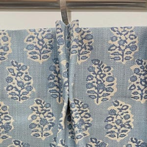 May include: Light blue fabric with a repeating floral pattern in white and darker blue. The fabric is draped, showing the texture and design of the material. The floral pattern is a stylized botanical design.