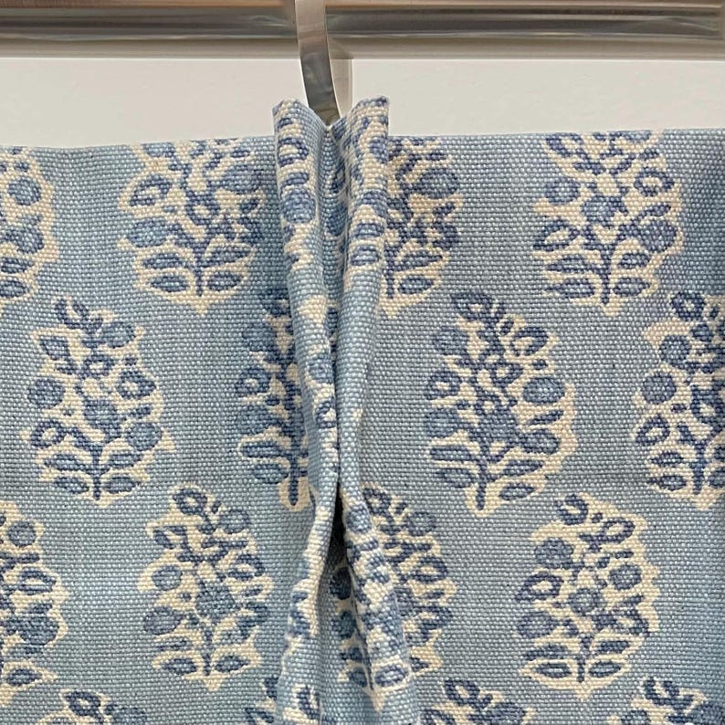 May include: Light blue fabric with a repeating floral pattern in white and darker blue. The fabric is draped, showing the texture and design of the material. The floral pattern is a stylized botanical design.