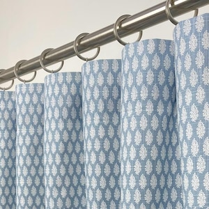 May include: Light blue and white patterned curtains hanging from a silver rod with silver rings. The curtains have a repeating white leaf design on a light blue background. The fabric is textured, and the curtains are partially closed.