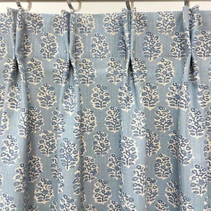 May include: Light blue curtain panel with a repeating floral pattern in white and darker blue. The fabric is pleated and hangs from silver metal rings on a rod. The design is a classic, elegant style.