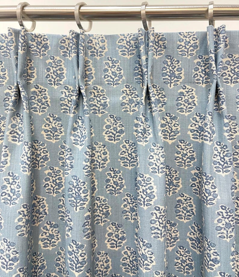 May include: Light blue curtain panel with a repeating floral pattern in white and darker blue. The fabric is pleated and hangs from silver metal rings on a rod. The design is a classic, elegant style.