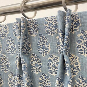 May include: Close-up of blue and white patterned curtains hanging from a silver rod. The curtains feature a repeating floral design in shades of blue and white. The fabric is textured, and the curtains are neatly pleated.