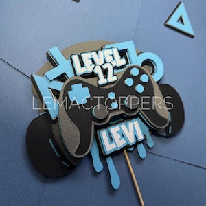 May include: A blue and black cake topper featuring a video game controller, the words "Level 12" and "Levi", and blue dripping paint.
