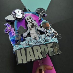 May include: A vibrant cake topper with a gaming theme, featuring a Marshmello character, a llama, and the name "HARPER" in silver lettering. The number "8" is also included. The design incorporates various game-related elements.