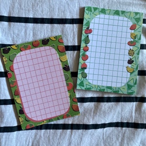 May include: Two green and white gridded notepads with a fruit border. The notepad on the left has a pink grid and a border of red, orange, yellow, and green fruit. The notepad on the right has a white grid and a border of red, green, yellow, and brown fruit.