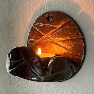 May include: A ceramic wall-mounted candle holder with a lit tea light. The holder is brown with a textured, geometric design on the backplate. The candle holder has a curved, leaf-like shape to hold the candle.