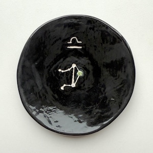 May include: A round, black ceramic plate with a glossy finish. The plate features a white Libra symbol and constellation design. A small, green accent is incorporated into the constellation. The plate is centered on a white background.