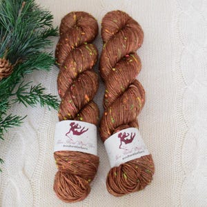 Può includere: Due gomitoli di filato tinto a mano in una base marrone con macchie multicolori. Il filato è avvolto su se stesso e ha un'etichetta che dice "Nina Hand Dyed Yarn #ninahanddyedyarn".
