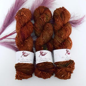 Puede incluir: Tres madejas de hilo teñido a mano en tonos marrón, óxido y verde, con etiquetas blancas con un logotipo y texto. El hilo está dispuesto sobre una superficie blanca con una ramita de hierba ornamental púrpura.