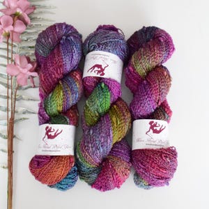 Puede incluir: Tres ovillos de hilo teñido a mano en tonos de morado, verde y naranja. El hilo es una mezcla de fibras y tiene un aspecto moteado. Los ovillos están etiquetados con "Nova Hand Dyed Yarn".