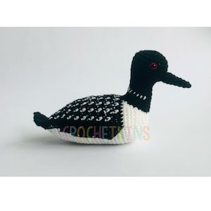 May include: A handmade crochet loon bird in black, white, and red. The bird has a black head and beak, a white chest, and a body with a black and white speckled pattern. The text "CROCHETAINS" is visible on the bottom.