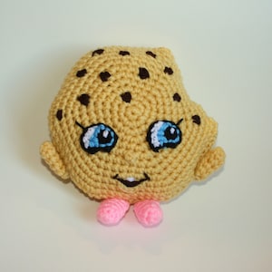 May include: A crocheted cookie plush toy with a smiling face, blue eyes, and pink feet. The cookie is tan with brown chocolate chips. The toy has small arms and is made of yarn.
