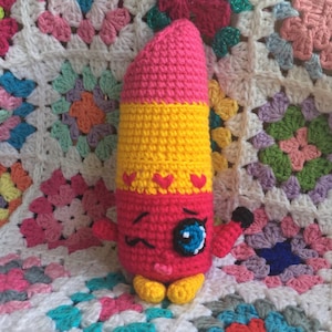 May include: A handmade crochet lipstick doll in pink, yellow, and red. The doll has a winking face with a blue eye, small arms, and yellow feet. The lipstick has a pink top, yellow middle, and red base with heart accents. The doll is set against a colorful crocheted blanket.