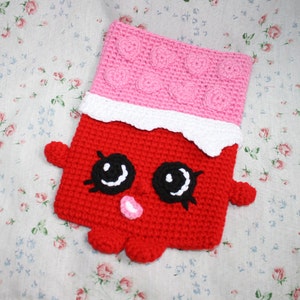 May include: A handmade, red crochet chocolate bar with a smiling face, black eyes, and a pink nose. The top section is pink with heart-shaped details, and a white, wavy edge separates the sections. The item is placed on a floral patterned fabric.