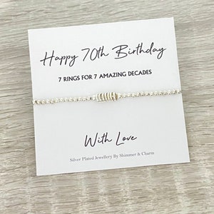May include: A silver bracelet with seven rings, a card that says "Happy 70th Birthday" and "7 rings for 7 amazing decades", and the words "With Love" and "Silver Plated Jewellery By Shimmer & Charm".