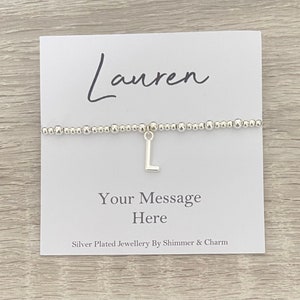 May include: Silver plated bracelet with a charm in the shape of the letter L. The bracelet is on a white card with the name "Lauren" written in cursive and the text "Your Message Here" and "Silver Plated Jewellery By Shimmer & Charm".