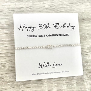 May include: A white card with the text "Happy 30th Birthday 3 Rings for 3 Amazing Decades" and "With Love" with a silver bracelet with three rings on it.