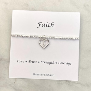Op de afbeelding: Een zilveren kralenarmband met een hartvormige bedel en een kruis. De armband wordt gepresenteerd op een witte kaart met het woord "Faith" en de woorden "Love, Trust, Strength, Courage". De merknaam "Shimmer & Charm" staat ook op de kaart.