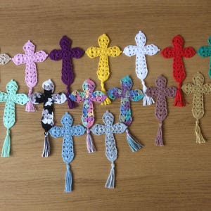 Könnte beinhalten: Eine Sammlung gehäkelter Kreuze in verschiedenen Farben, darunter Rosa, Lila, Gelb, Weiß, Rot und Grün. Jedes Kreuz hat eine Quaste am unteren Ende. Die Kreuze sind auf einer Holzoberfläche angeordnet.