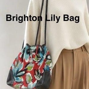 May include: A Brighton Lily Bag featuring a red, blue, and green floral pattern with dark brown leather accents and braided shoulder straps. The bag has a drawstring closure with a silver-toned metal charm attached to the front cord.