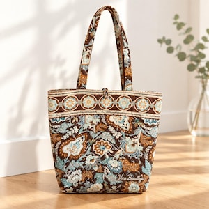 May include: A quilted tote bag featuring a brown, light blue, and cream floral paisley pattern. The bag has two shoulder straps, a top border with a circular motif, and the text Vera Bradley embroidered in white on the lower front section.